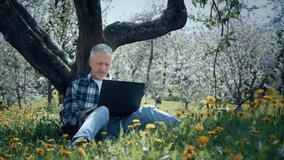 An elderly white-haired man is working on a laptop, sitting on the grass among the yellow flowers in the park under a tree on a sunny summer day. - Powered by Shutterstock - Get 15% off with code: PIKWIZARD15