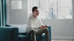 Delighted brunet guy in white hoodie plays online video game holding wireless joystick and smiles sitting at home slow motion