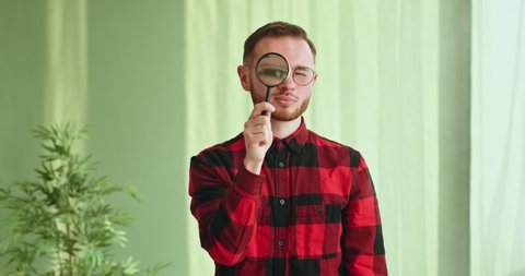 Funny young guy in glasses and a checkered shirt looking at the camera through a magnifying glass magnifying glass researcher scientist standing indoors funny video