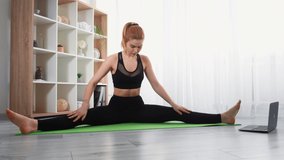 Sport at home. Internet workout. Morning fitness. Athletic woman in sportswear watching video training on laptop doing stretching exercise on yoga mat at light modern interior.