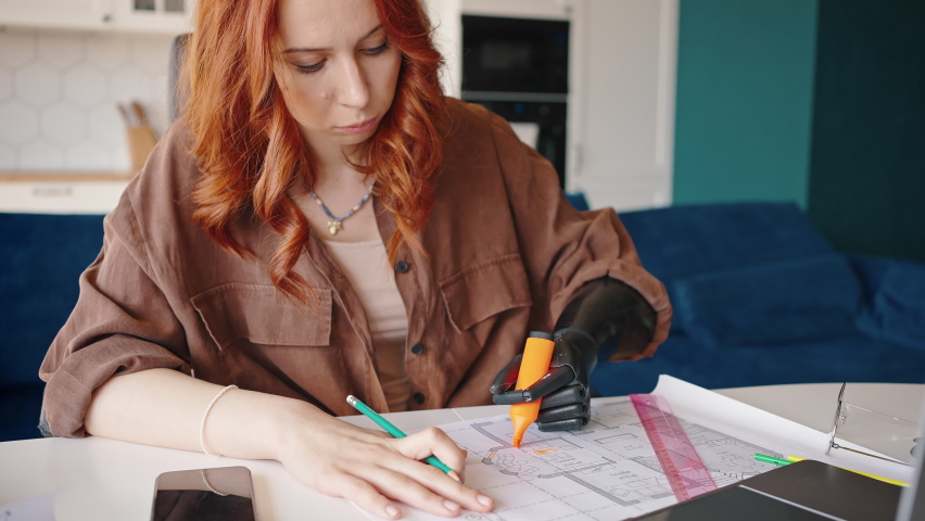 Red-haired attractive girl designer with a prosthetic limb is working on a project in her home office. Marks up drawings with a pencil and marker. Royalty-Free Stock Footage #1074611459