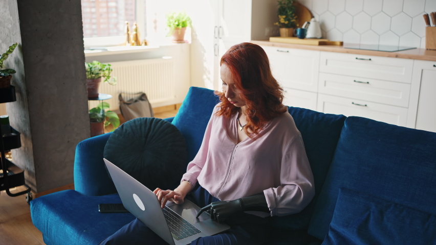 Woman deftly controls the touchscreen of the laptop with the fingers of the prosthesis. Happy ginger girl with artificial limb working with a laptop at home. Royalty-Free Stock Footage #1074611465