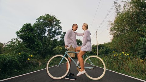 Biking road trip. Love couple on blue bike in white clothes on forest road. Just married woman and man kiss, hugs, stand on bicycle. Cycle Fix.
