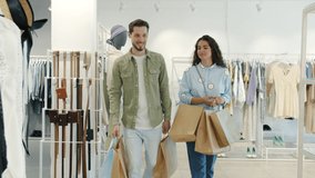 Slow motion of happy young couple walking in shop holding bags talking and smiling looking at clothes on hangers. Relationship and shopping concept. - Powered by Shutterstock - Get 15% off with code: PIKWIZARD15