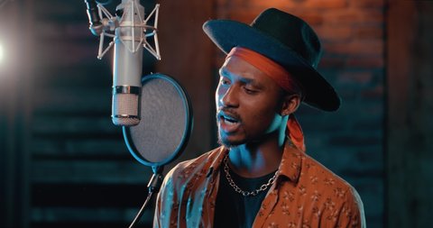 Stylish Afro-American Singer Recording Song with a Microphone in Music Studio. Professional Rap Performer wears bandana and accessories Actively Singing new Composition. Colorful Recording Studio.