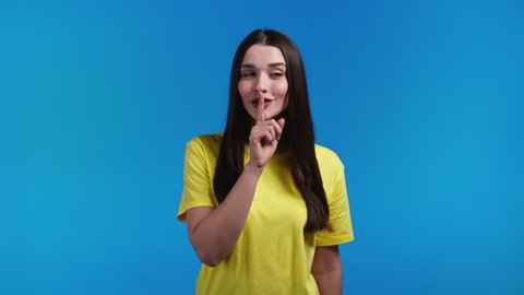 Smiling woman holding finger on her lips over blue background. Gesture of shhh, secret, silence. Close up