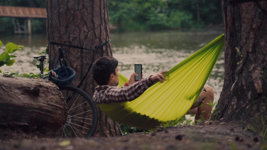 Man traveling on bicycle resting in green hammock, surfing Internet on smartphone in forest near lake. Cyclist in hammock in park by river uses phone. Male came to forest on bike and relax in hammock