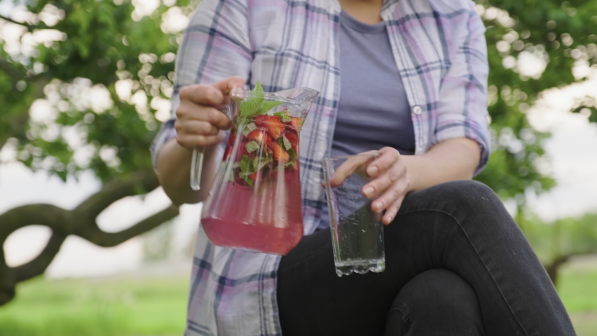 Close-up of female hands with jug pouring fresh natural drink with strawberries and mint into glass.