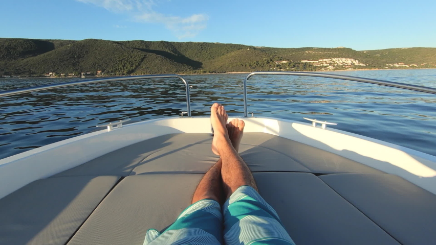 Slow motion - POV of tan man enjoying ride on a motor boat at sunset. First person view of crossed male legs on the bow of a speed boat. Young adult boy having fun cruising on the sea | Shutterstock HD Video #1074632003