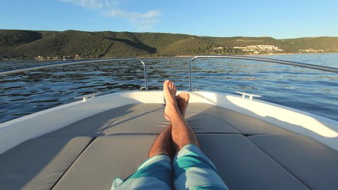 Slow motion - POV of tan man enjoying ride on a motor boat at sunset. First person view of crossed male legs on the bow of a speed boat. Young adult boy having fun cruising on the sea