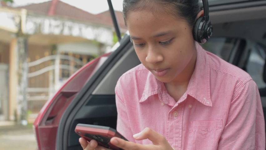 Asian female teenager watching social media or playing games online from smartphone with headphones and sitting at rear side of hatchback car while waiting parents driving her to school.