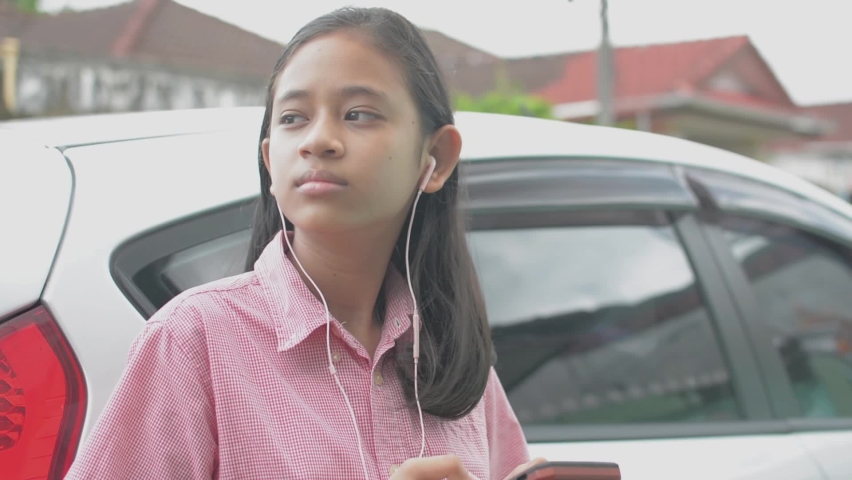 Adorable long hair Asian high school girl using smartphone for social online communication with earphones and standing next to a car while waiting parents driving her to school. Teenager lifestyle.