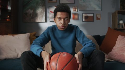 Serious black man teenager holding basketball ball and sitting on sofa in living room, young teen athlete, sport portrait at home, high school kid preparing for watching professional game on tv.