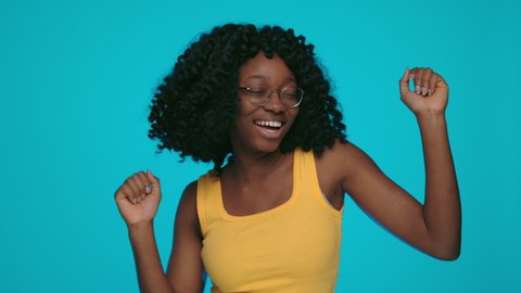 Carefree young woman with dark curly hair enjoying disco time over yellow background. Happy african female in eyeglasses dancing and singing at studio.