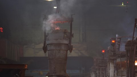 The melting bowl with the molten metal moves along the crane beam in the steel production, smoke and fire from the crucible.