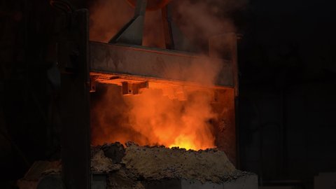 The melting bowl with the molten metal moves along the crane beam in the steel production, smoke and fire from the crucible, close-up.