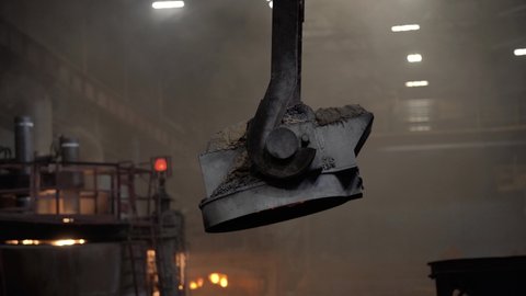 A bowl of molten metal moves along a crane beam in the steel industry, smoke and fire from the crucible, the general plan.