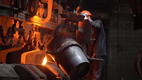A steelworker pours molten metal from a crucible into molds in the steel industry, a conveyor moves.