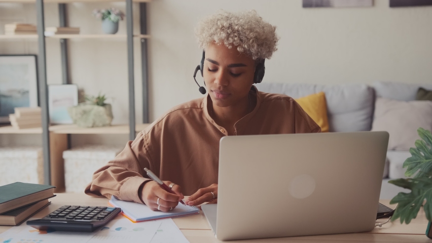 Young African business woman call center agent at home workplace, cheerful dark skinned female telemarketer operator wear headset having work in customer service support office make notes calculation. Royalty-Free Stock Footage #1074652691