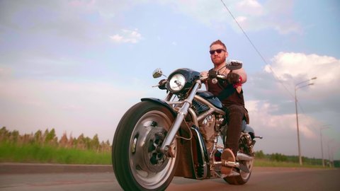 Stylish biker with tattoos rides a motorcycle on a country road at sunset