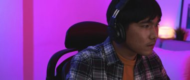 Young Asian man gamer wearing headphones focusing on playing e-sports in front of computer screen at night 