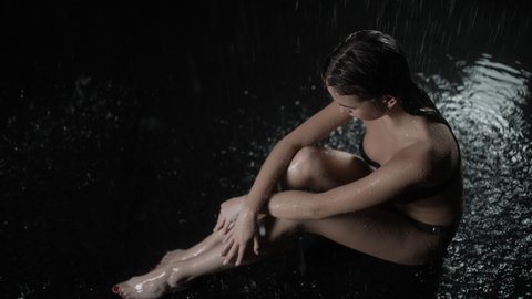 Attractive young millennial woman relaxing under running water splashes. Wet woman in black swimsuit with slim beautiful fit body under the falling drops, stream of water, beauty and body care concept.