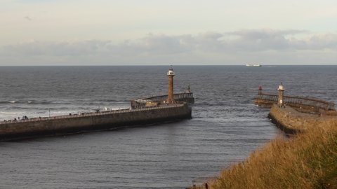 Whitby town sea breakwaters with lighthouse and ship sailing past in the sea near horizon before sunset.