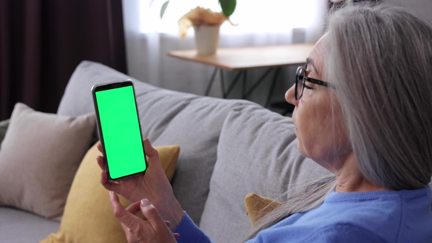 Elderly gray-haired woman making video call at smartphone camera green screen. Senior lady talking and listening during video chat with friends or colleagues. Over shoulder mobile phone screen view.