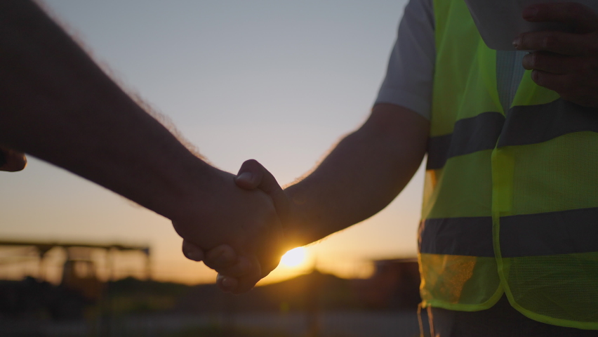 Portrait of hands of two builders. Builder shaking hand the builder on built house background. Close up of a handshake of two men in green signal vests against the background of the sun