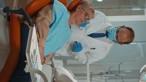 Vertical video Stomatologist performing examination and taking care of teeth using dental tools. Orthodontist speaking to woman sitting on stomatological chair while nurse preparing for surgery in