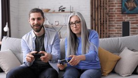 Adult son and senior mother enjoy spending time together. Grandmother and grandson playing video games using gamepads at home. Mothers Day. Two generation family. Active modern elderly people.