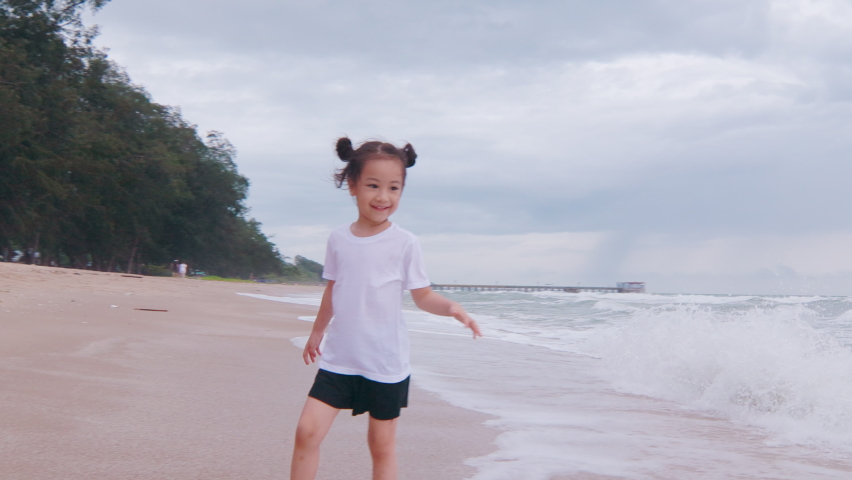 4K of adorable 5 years old asian girl with black hair is running with happy smile and enjoyment near ocean coast seeing strong wave and horizon for sunset which is relaxing holiday for kid and family.