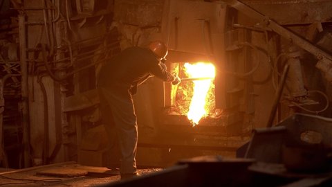 A steelworker clears a blast furnace with a metal scrap, molten metal flows, a smelter, smoke and fire.