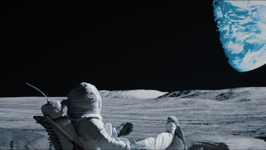 Astronaut sits in a beach chair on a Moon surface, holding phone in hands. Shot with 2x anamorphic lens | Shutterstock HD Video #1074671849