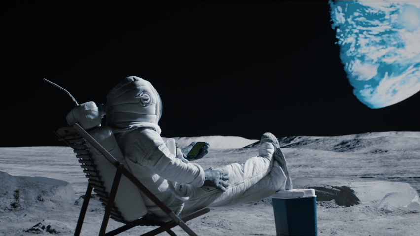 Astronaut sits in a beach chair on a Moon surface, holding phone in hands. Shot with 2x anamorphic lens Royalty-Free Stock Footage #1074671849