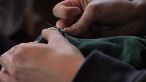 Female hands hand sew green jacket material with thin metal needle and string thread to make alteration and tailoring adjustment, side perspective macro close up view pulling thread through.