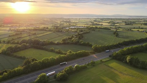 Aerial View Of Vehicles Driving At M5 Motorway Passing By Green Fields At Dursley Town At Sunrise In England, UK.