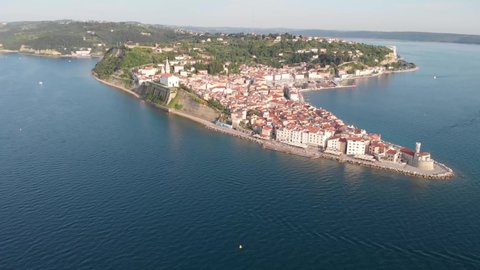 Ancient buildings with red roofs and Adriatic sea. Drone clip of beautiful peninsula Piran, Slovenia in the sunset. Church on top of the cliff and fortress on the end of the land.