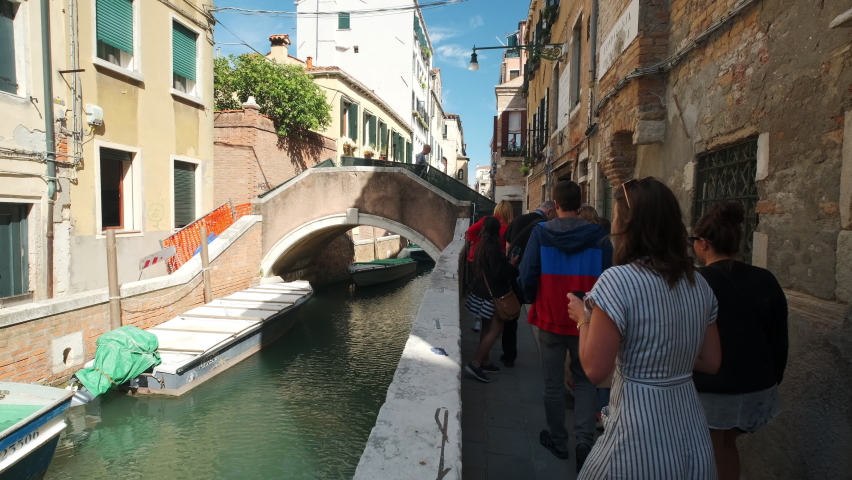 Venice , Italy - 09 18 2019: Slow motion pov showing group of tourist walking along narrow italian alleys beside famous Venetian canal during sightseeing tour. Beautiful old architecture housing areas