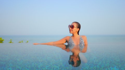 Portrait shot of Thai woman in sunglasses resting inside infinity pool leaning on the arm on the edge of pool, face reflection in water surface daytime, endless sea on background