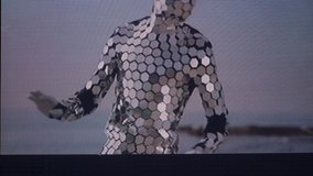 A man in a full body disco suit dancing next the sea in the morning sunlight with overlayed video distortion effects