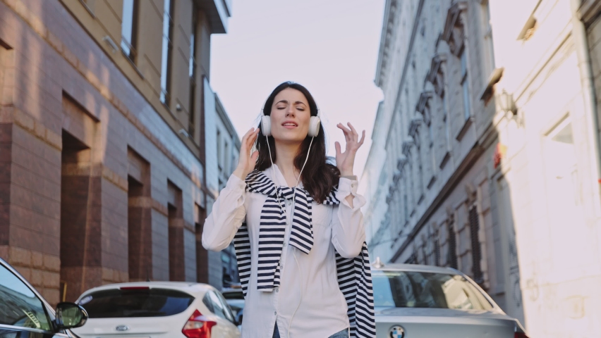 Dynamic caucasian woman wearing casual clothes and headphones listening to music and enjoying rhythm gesturing while dancing at the street. Teen people concept