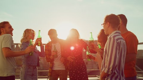 Beautiful people toasting with drinks at sunset rooftop party. Multiethnic friends clinking with beer bottles on terrace in city background. Smiling men and women enjoying time together outdoors. 