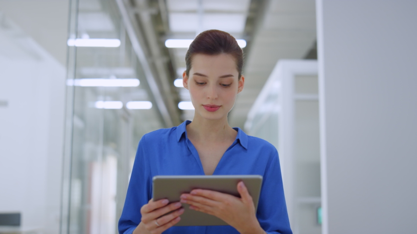 Beautiful businesswoman looking tablet screen while walking on modern office. Happy woman reading message on digital device indoors. Joyful business lady working while going along creative space.