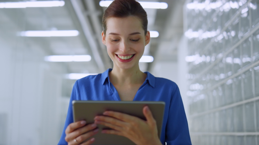 Beautiful businesswoman looking tablet screen while walking on modern office. Happy woman reading message on digital device indoors. Joyful business lady working while going along creative space.