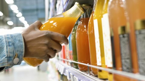 Black man in blue denim jacket hand takes orange natural juice from large grey metal shelf in local supermarket close view