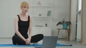 Adult middle aged athletic woman trainer recording online yoga class broadcasting. 40s female looks at laptop sitting on floor trains with coach video chat conference folds palms in front of her bows