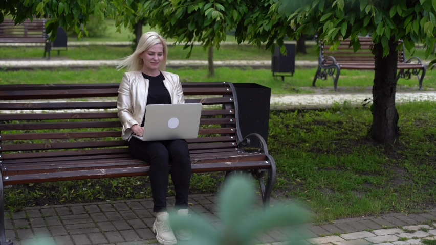 Caucasian woman in headset typing on the laptop computer on the bench in the park. Female student makes conference video call on laptop computer talks with web tutor, online teacher in webcam chat.