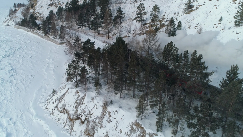 Aerial forward beutiful old steam locomotive, engine rides on railroad railway, releases thick white clouds of steam from pipe. Winter snowy nature. Historical Soviet Union train Circum-Baikal Russia