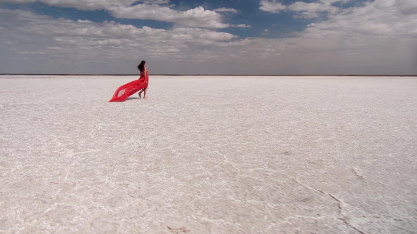 Flight around beautiful young girl woman model in red dress fluttering in wind walks along dry desert cracked bottom of salt mineral lake. Amazing alien landscape. Fashion, beauty. For advertising